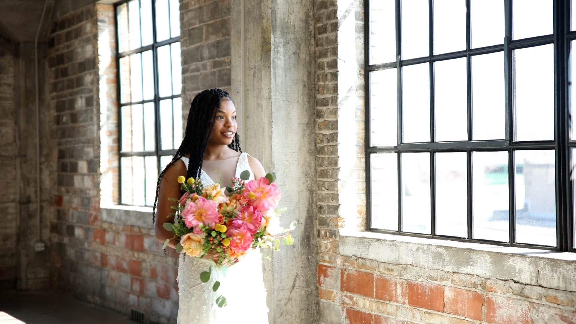 A bride stands in a brick building holding bright pink and yellow flower bouquet looking out a window at one of the best minneapolis wedding venues
