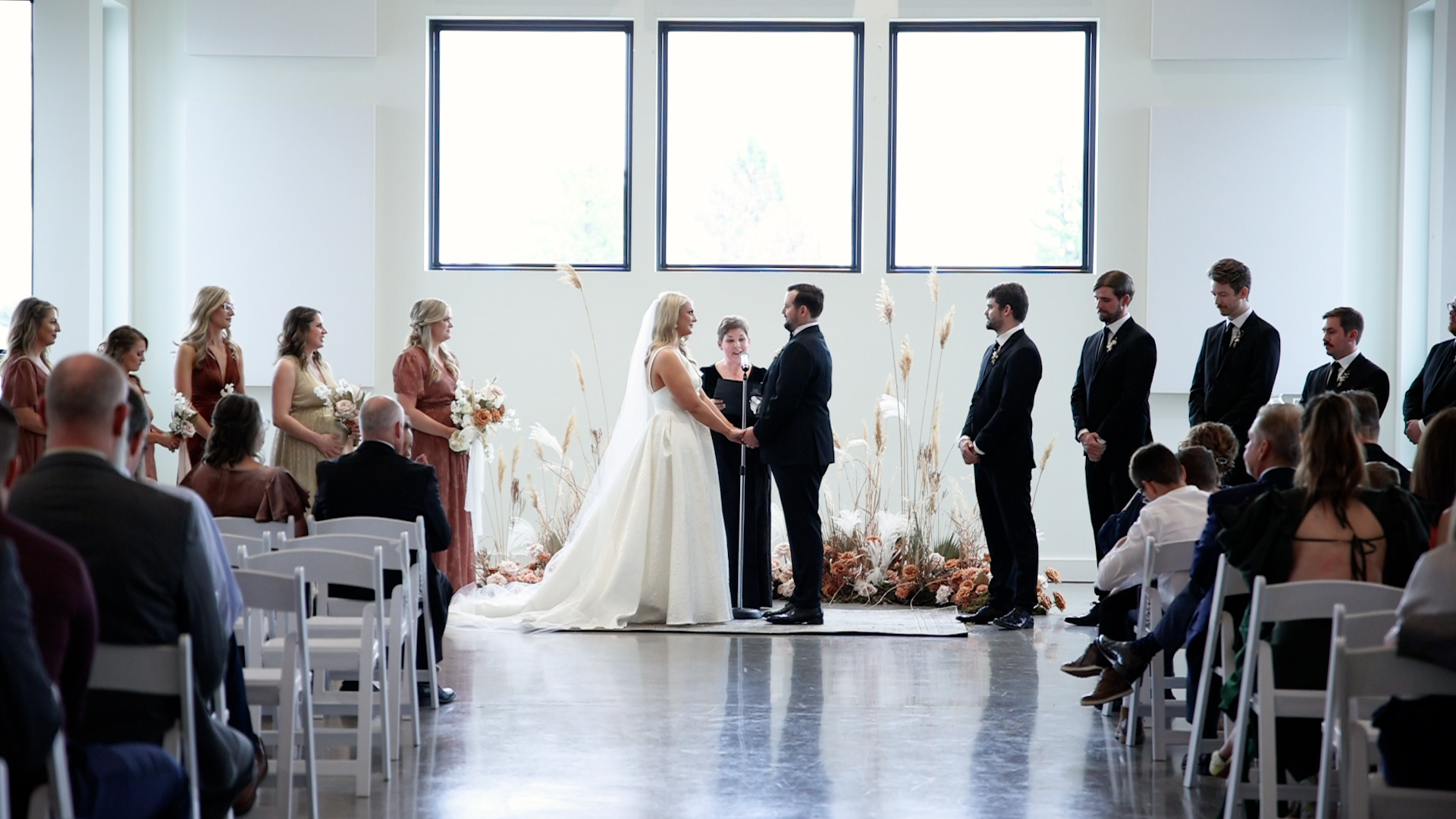 A bride and groom stand at their ceremony holding hands in front of three large windows Woodhaven weddings