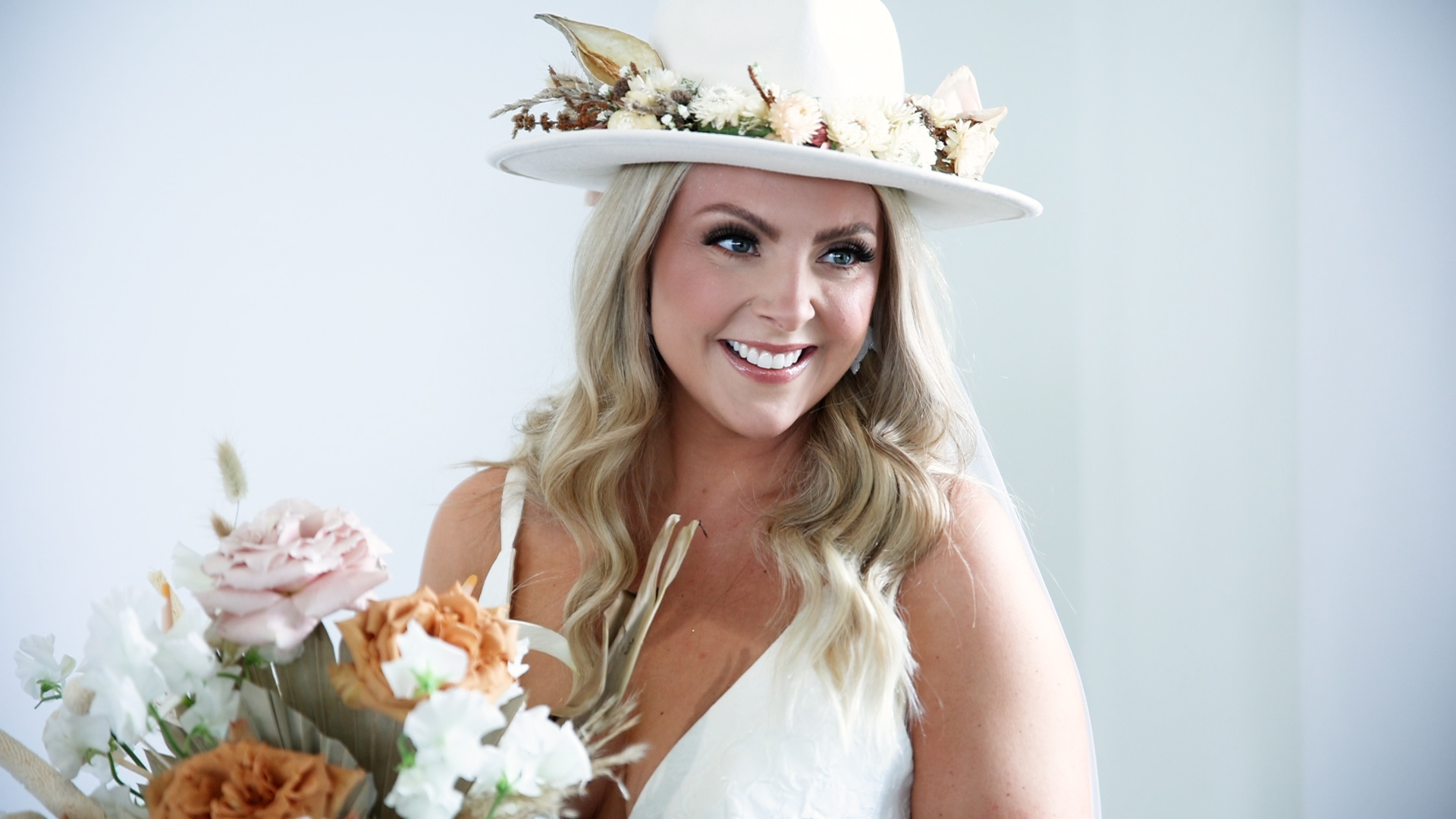 A bride stands by a window in a white hat decorated with florals while holding her matching bouquet