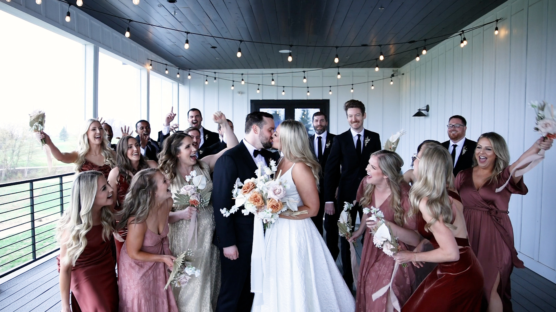 Newlyweds kiss under string lights on a porch while the wedding party celebrates around them Woodhaven weddings